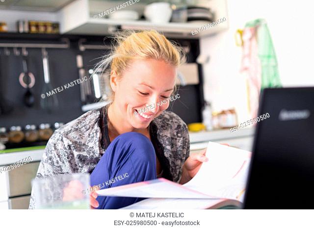 Female student in her casual home clothing working and studying remotly from her small flat in the morning. Home kitchen in the background