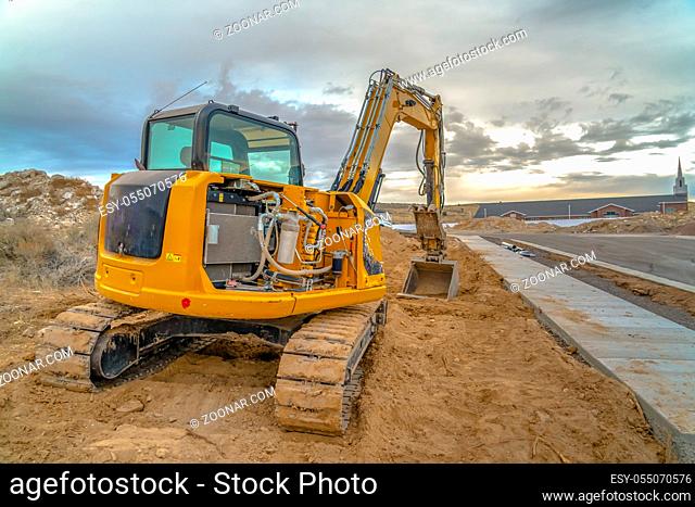 Excavator with Utah Valley church in background. Excavator on dirt beside a road on a construction area in Utah Valley. A church and glowing sky with clouds can...