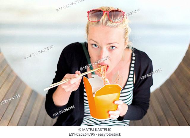 Beautiful young woman, sitting on the bench in park, holding a fast food lunch box, eating noodles from Chinese take-away with traditional wooden chopsticks