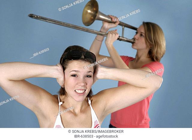 Girl practicing her trombone while her friend covers her ears, annoyed