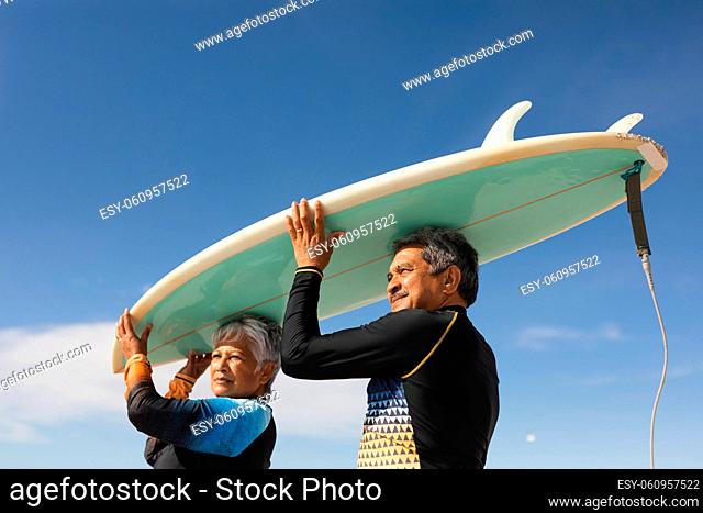 Low angle view of multiracial senior couple carrying surfboard over heads against blue sky