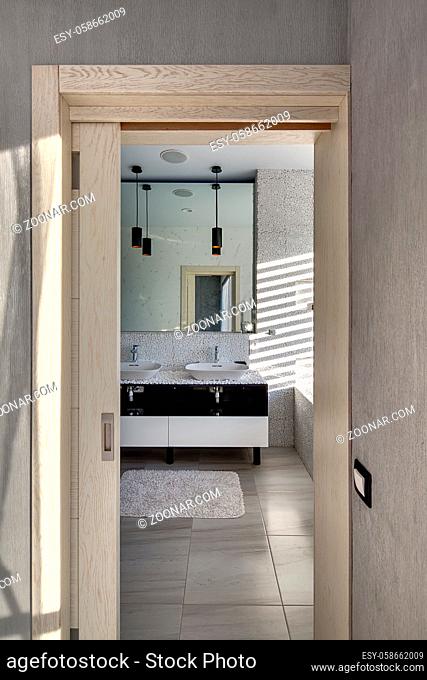 Entrance to the bathroom in a modern style which decorated with the pebbles. There is a large mirror, two sinks with faucets and lockers under them