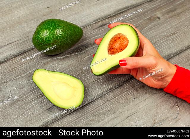 Woman hand holding avocado cut to half, with other whole one on gray wood table in bacgkground