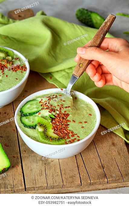 Freshly prepared smoothies. A woman's hand with a spoon takes a green smoothie of cucumber, avocado, kiwi and flax seeds against a wooden board