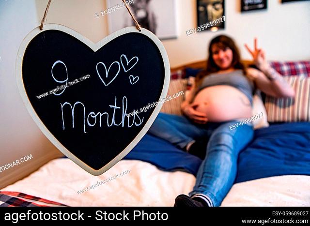 A shabby chic heart blackboard sign is seen hanging in a bedroom with message nine months, as a heavily pregnant woman lies in bed with shallow depth of field