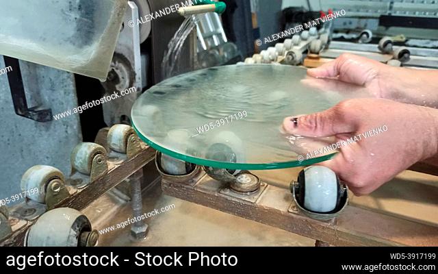 Worker hands grind glass on a machine. Jets of water cooling machine processing of glass. Glass blank working on grinding machine