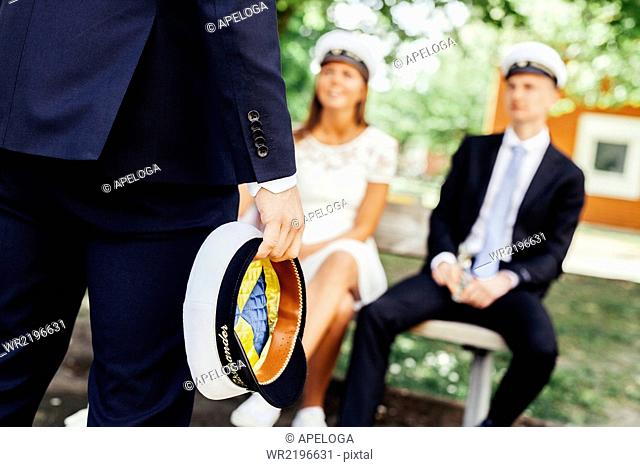 Midsection of graduate holding cap while friends sitting on bench at party
