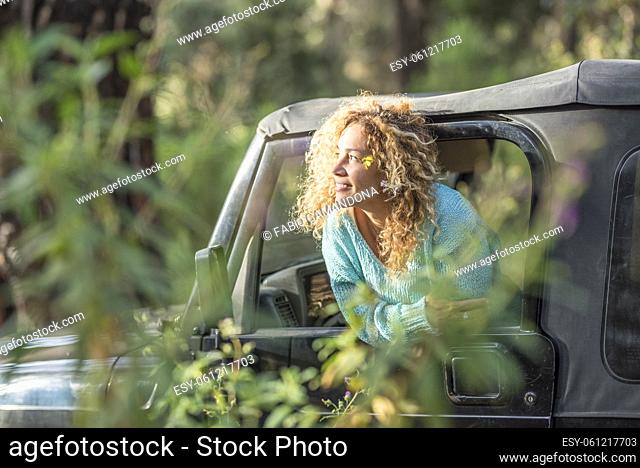 Young woman leaning outside car window and looking at forest. Beautiful woman enjoying holiday trip in forest. Happy woman exploring forest and admiring nature...