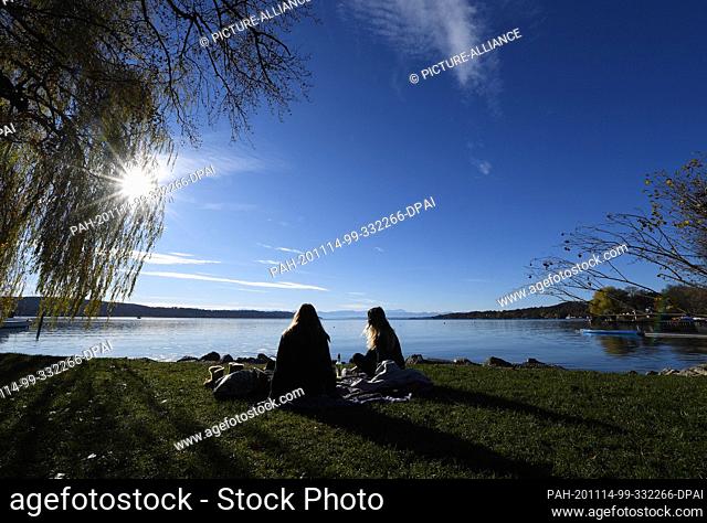 14 November 2020, Bavaria, Starnberg: Two young women are sitting in the sun at Lake Starnberg, in the background the Wetterstein mountain range with the...
