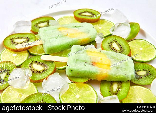 Close-up of healthy home-made fruit ice cream on a stick with a slice of peach in a plate with pieces of lime, kiwi and cold ice cubes