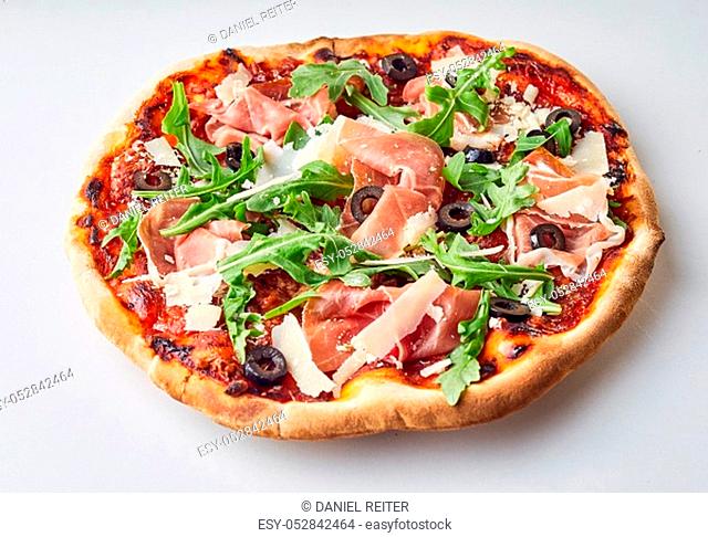 Traditional flame grilled crispy Italian pizza on a thin base topped with prosciutto, black olives, parmesan cheese and fresh rocket over a white background