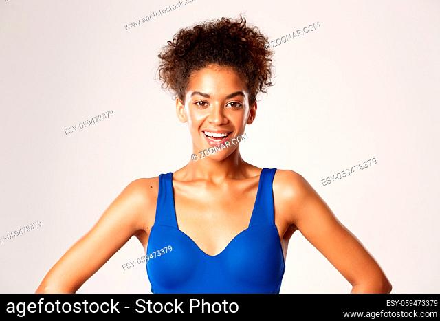 Close-up of attractive healthy woman in blue sports bra, smiling at camera, standing over white background
