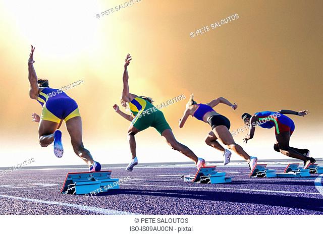 Four female athletes on athletics track, leaving starting blocks, rear view