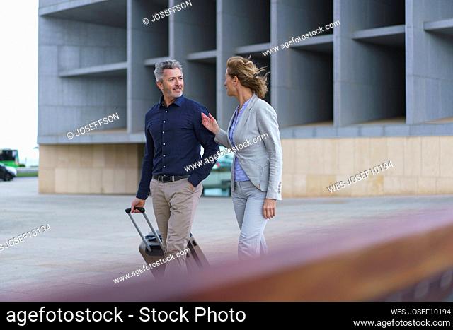 Smiling businessman with suitcase walking by businesswoman in front of office building