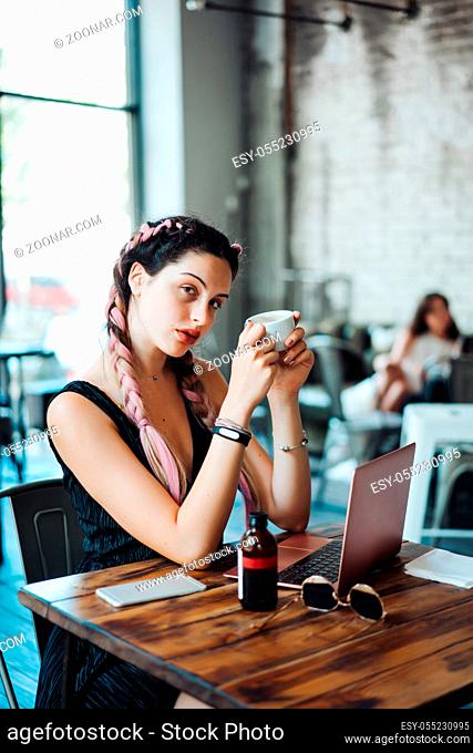 Young woman sitting in coffee shop at wooden table. On table is laptop, drinking coffee