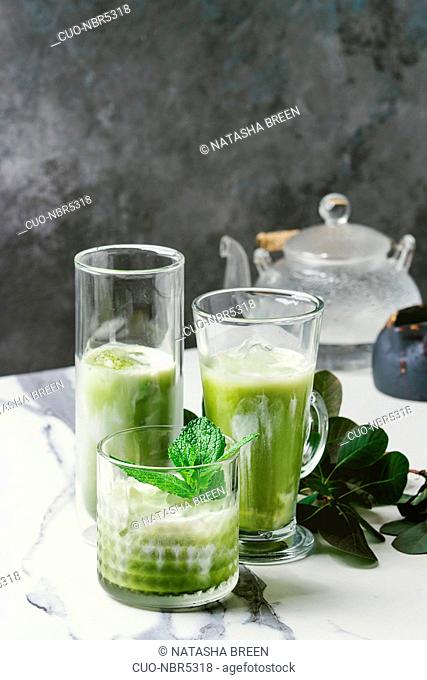 Matcha green tea iced latte or cocktail in three different glasses with ice cubes, matcha powder and jug of milk on white marble table