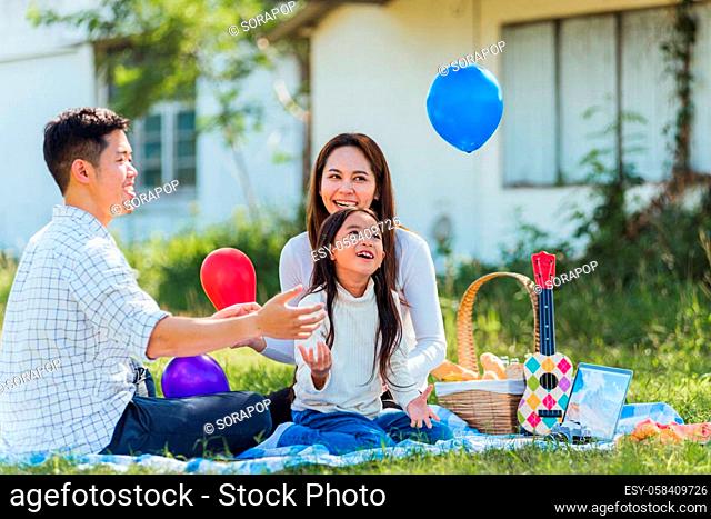 Happy Asian young family father, mother and child little girl having fun and enjoying outdoor sitting on picnic blanket playing balloons at summer garden spring...