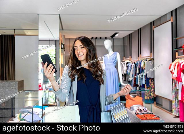Smiling woman looking at smart phone while carrying shopping bags in boutique