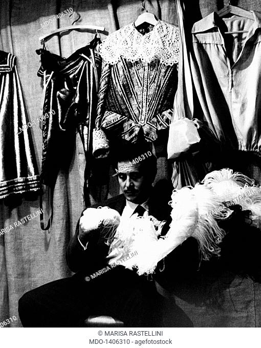 Domenico Modugno in his dressing room. The italian singer Domenico Modugno in his dressing room among some costumes; the artist is seated and has a feathered...