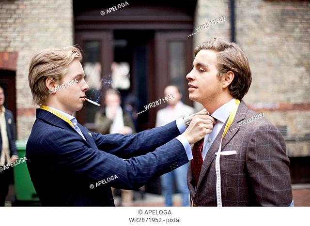 Handsome owner adjusting partner's necktie while smoking cigarette against building