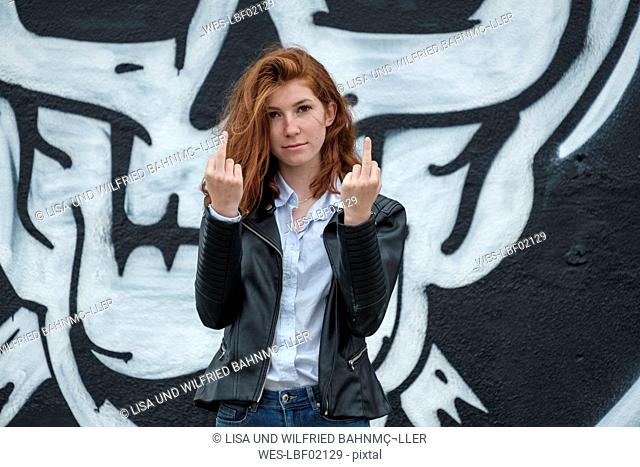 Italy, Finale Ligure, portrait of redheaded teenage girl showing the finger in front of wall with skull graffito