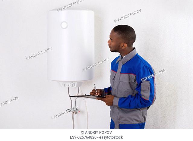 Young African Male Plumber Holding Clipboard Looking At Electric Boiler