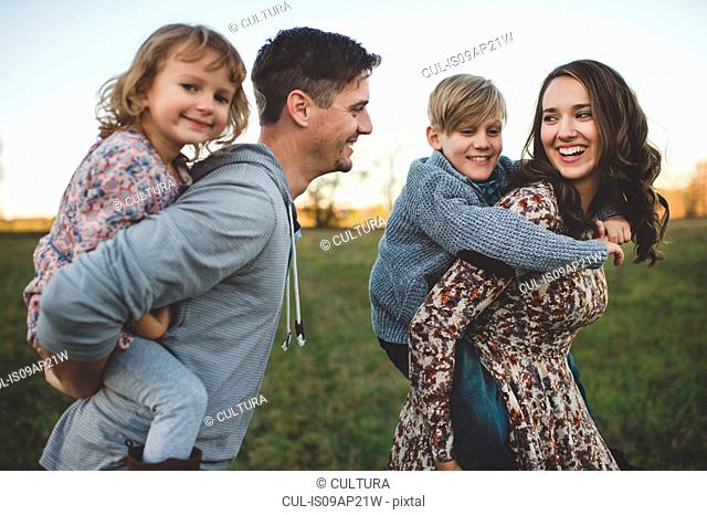 Young couple giving son and daughter piggy backs in field