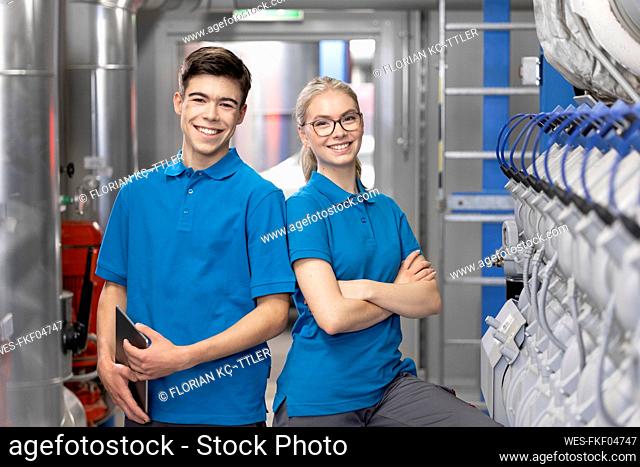 Smiling technician with arms crossed standing by colleague holding tablet PC in factory