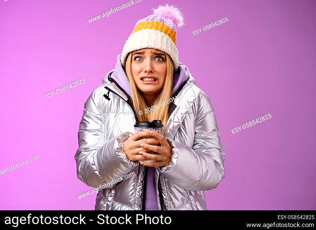 Shaking uncomfortable displeased girl wearing stylish silver jacket hat trembling freezing cold warm hands holding take-away coffee cup clench teeth pop eyes...