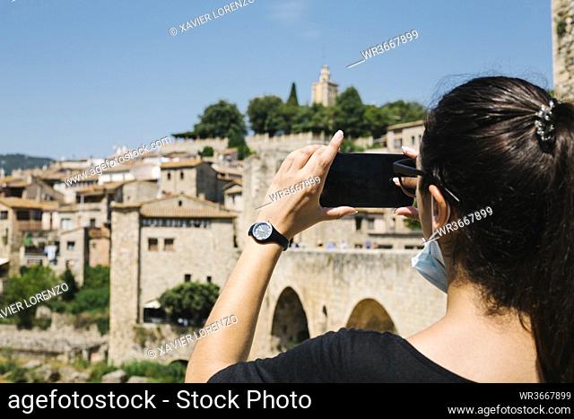 Woman wearing protective mask and taking a photo with her smartphone of Besalu, Catalonia, Spain