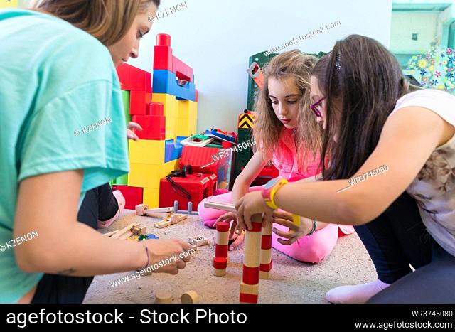 pre-school girls playing together with wooden toy blocks on the floor during playtime supervised by a careful young kindergarten teacher
