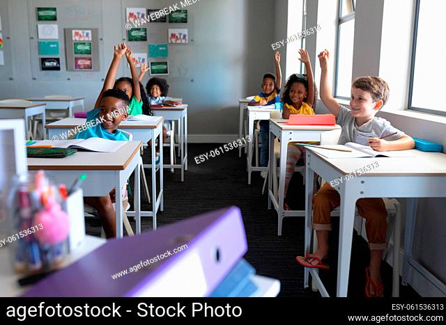Smiling multiracial elementary school students with hands raised sitting at desk in classroom