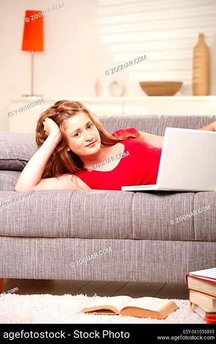 Smiling teen girl with laptop computer lying on couch at home looking at camera