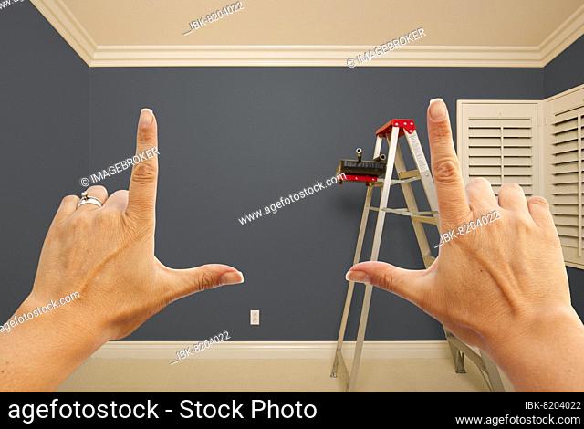 Hands framing grey painted room wall interior with ladder, paint bucket and rollers