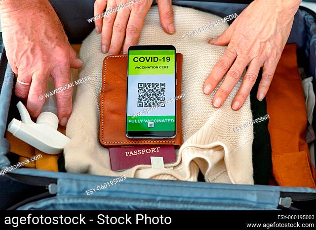 Hands of caucasian senior couple packing smartphone with covid vaccine passport into suitcase