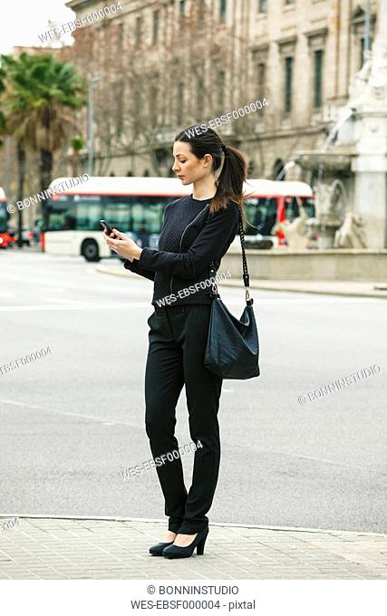 Spain, Catalunya, Barcelona, young black dressed businesswoman looking at her smartphone in front of a street