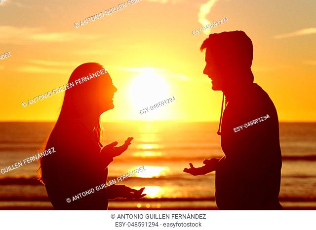 Side view of two friends or couple silhouette of teens talking happy on the beach at sunset with the sun in the middle and a warmth light