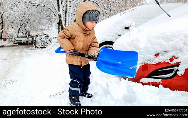 Boy in beige jacket and grey hat helping to clean up the snow covered red car after snow storm using big blue shovel