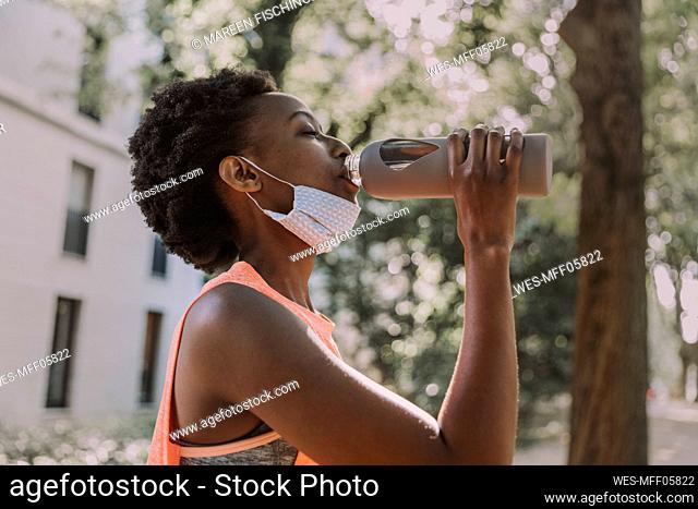 Sportive young woman with protective mask drinking water from flask