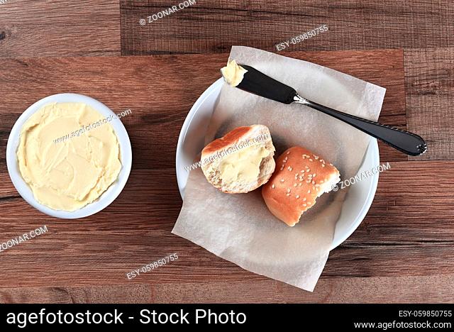 High angle view of a sesame dinner rolls broken in half and buttered. Next to the bread plate is a crock of butter with knife