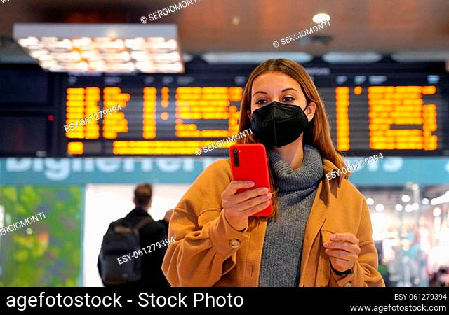 Traveler woman wearing KN95 FFP2 face mask at the subaway station. Young woman with behind timetables of departures arrivals checking information on her mobile...