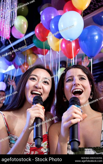 Two friends holding microphones and singing together at karaoke, balloons in the background