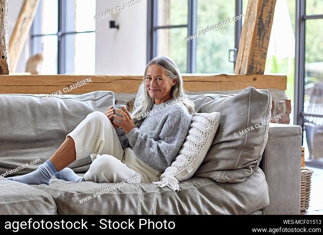 Smiling mature woman holding coffee cup while sitting on sofa at home