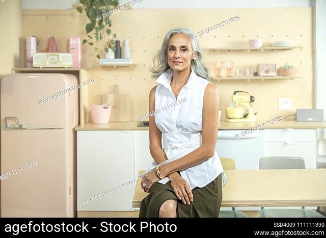 Wide interior shot in kitchen of middle-aged woman sitting with her legs crossed looking into camera