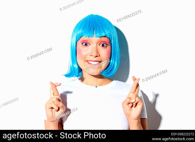 Excited beautiful asian girl in blue wig biting lip and looking hopeful at camera, making wish with fingers crossed, standing in halloween costume over white...