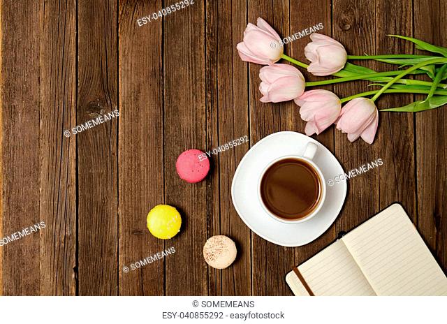 Cup of coffee, macarons, pink tulips and notebook on wooden background. Top view