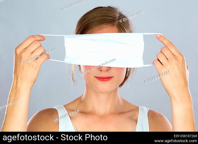Blond woman holding face mask covering her eyes in front of her head. Concept of hiding in fear of respiratory infection