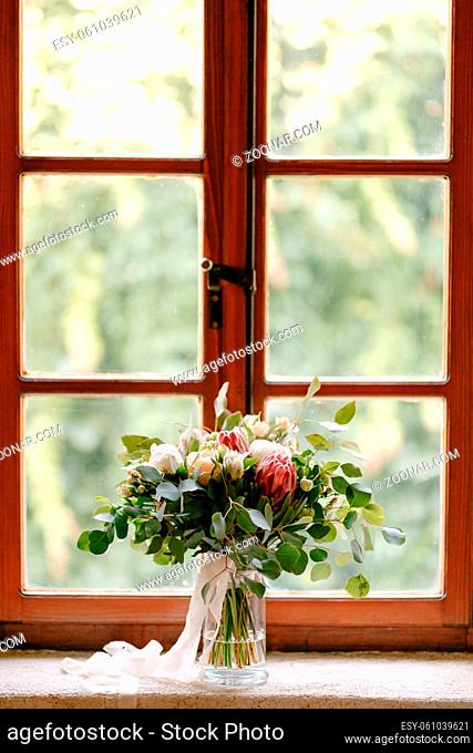 bridal bouquet of white peonies, roses, pink protea, snowberry, branches of eucalypt tree and white ribbons on the window. High quality photo