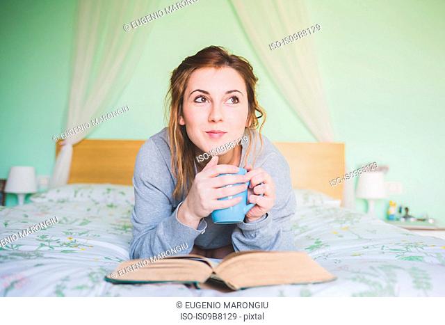 Young woman lying on bed holding coffee cup and looking up