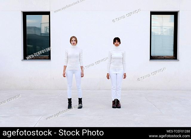 Young women maintaining social distance while standing against white building during coronavirus crisis